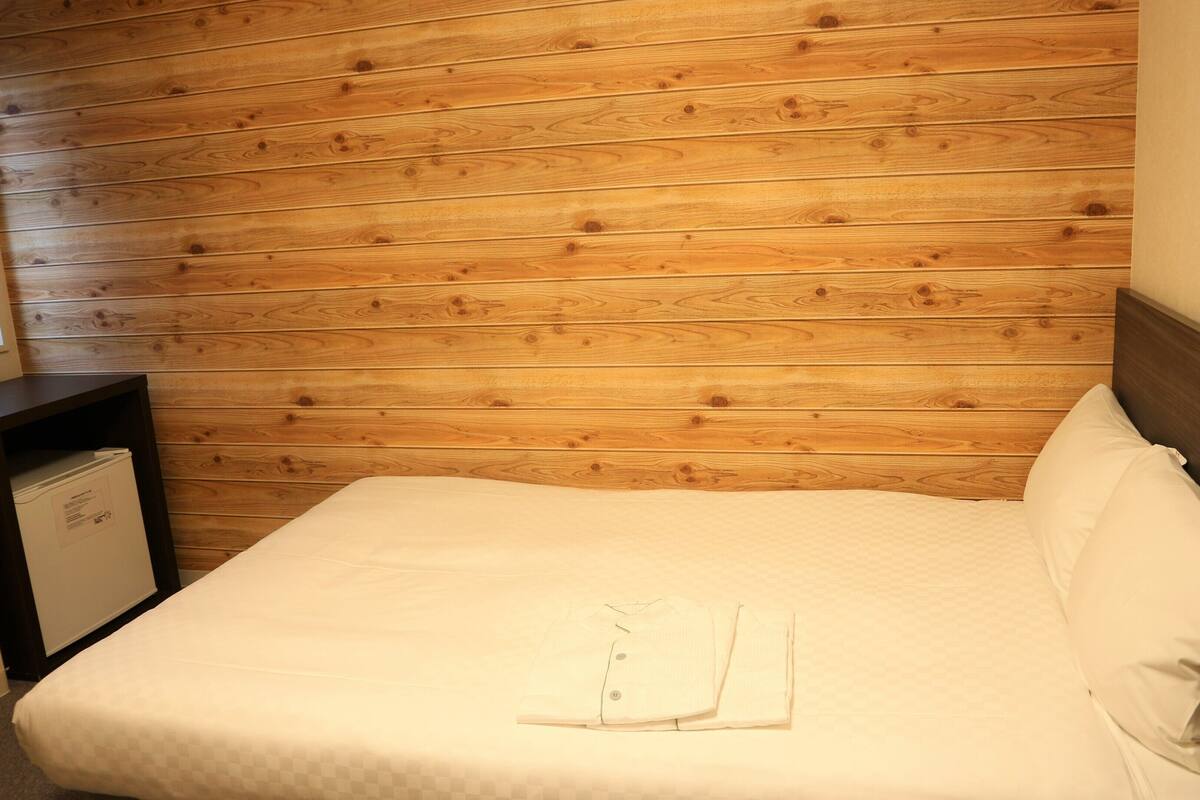 Una cama bien hecha con una camisa blanca doblada encima, paneles de madera en la pared y una mesita de noche con una lámpara.
