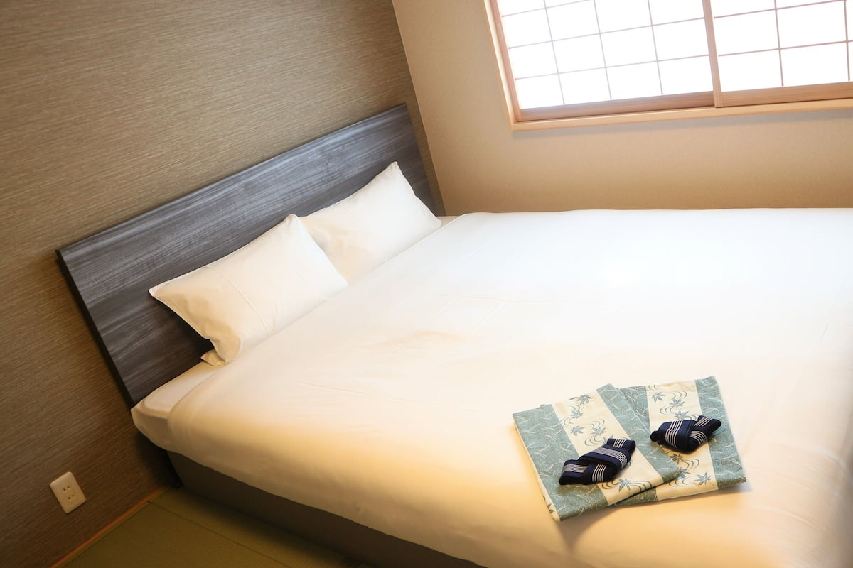Una cama bien hecha con sábanas blancas, cabecera de madera y una ventana con persianas.