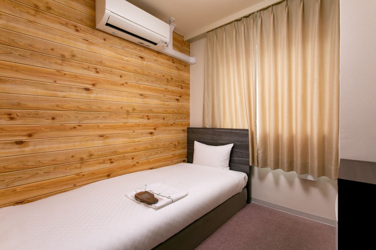 Una cama individual con cabecera de madera, almohada blanca y una mesita de noche con un libro y gafas.