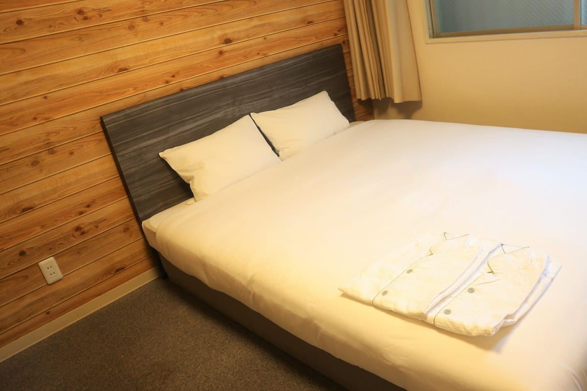 Una cama bien tendida con sábanas blancas y una toalla doblada, cabecera de madera y suelo alfombrado.
