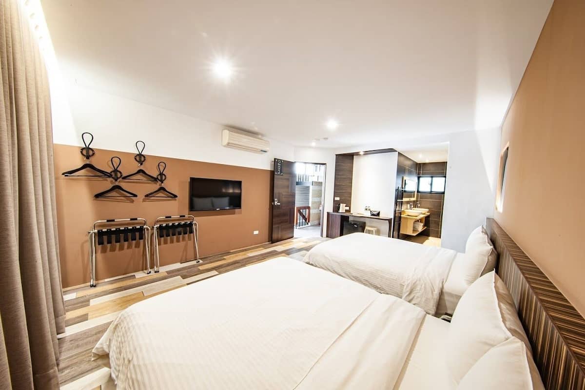 A hotel room with two beds, a TV, a desk with a chair, a bathroom, and a coat rack with hangers.