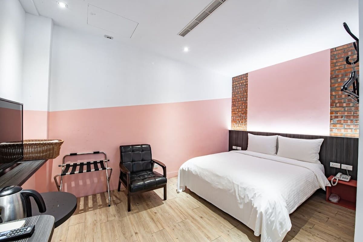 A hotel room with a bed, a desk with a kettle, a chair, and a basket.