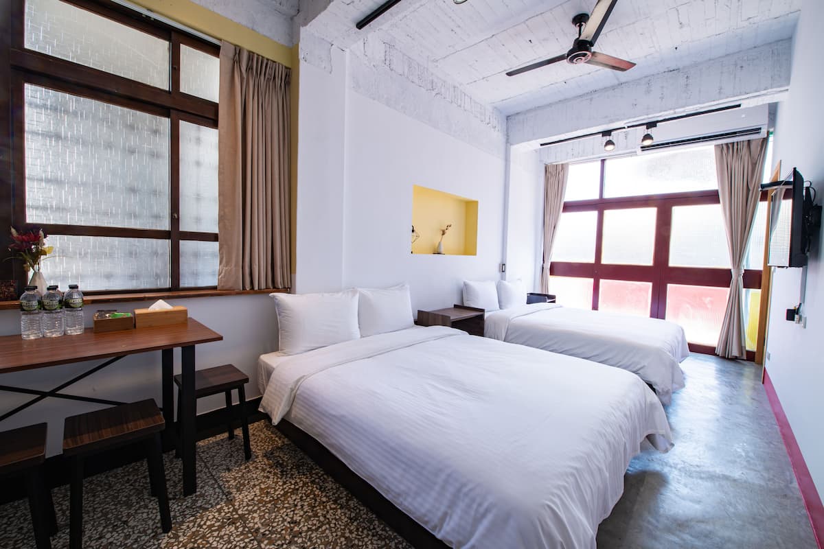 A hotel room with two beds, a desk with a chair, a small table with bottles, and a ceiling fan.