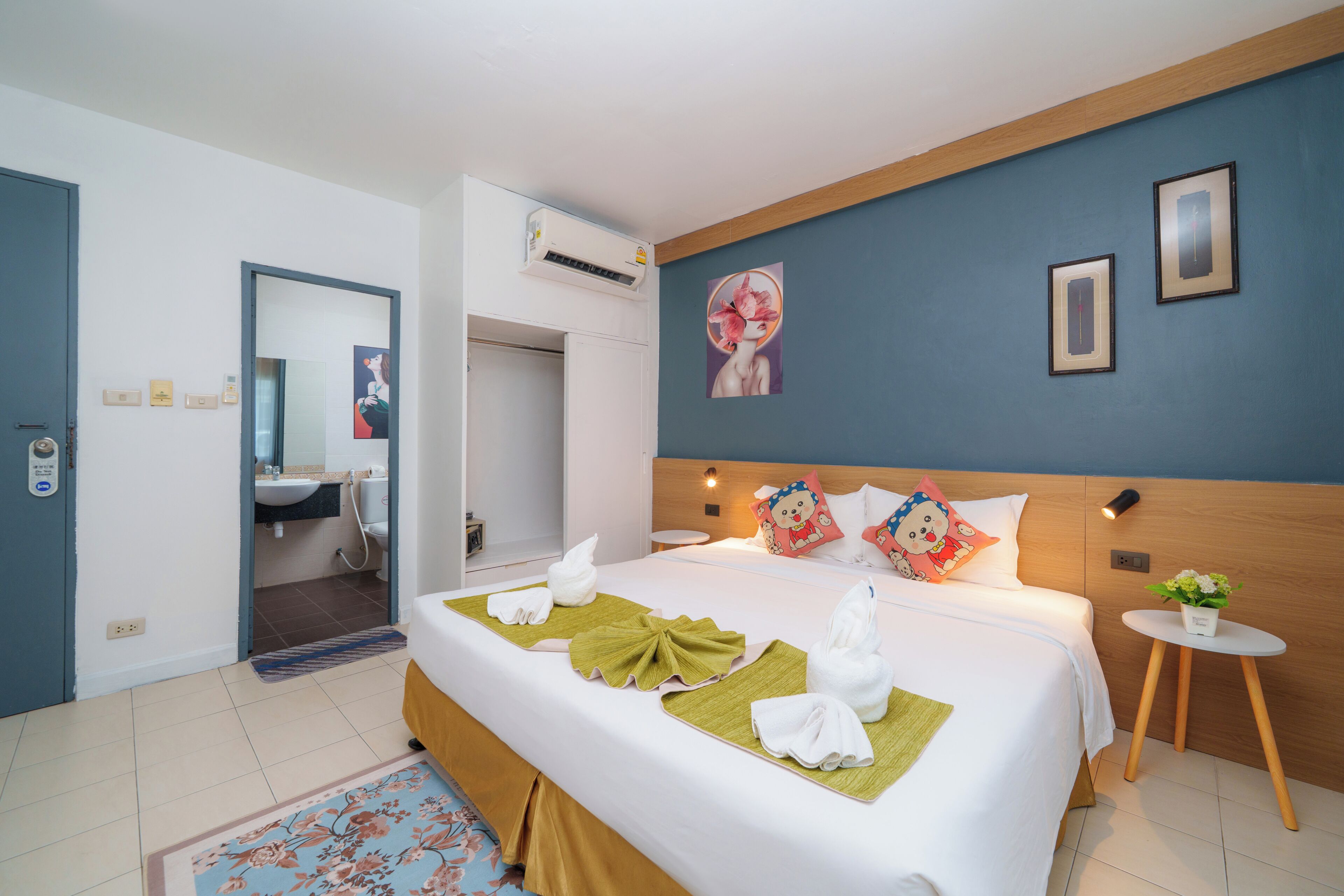 A hotel room with a bed, a small bedside table, a wall-mounted air conditioner, a mirror, and a bathroom visible through an open door.