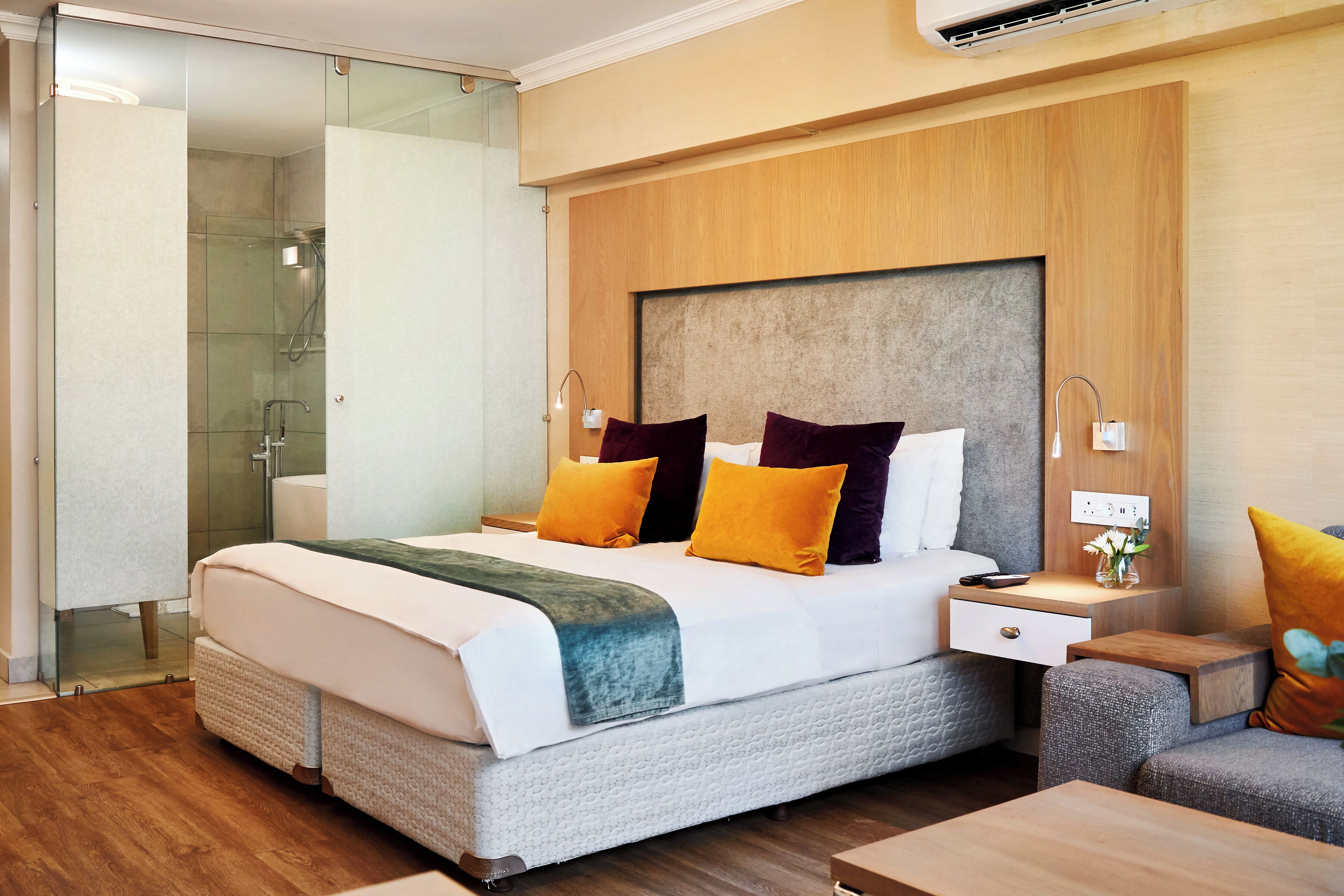A modern hotel room with a large bed, bedside tables, a nightstand, a glass shower enclosure, and a wooden headboard.