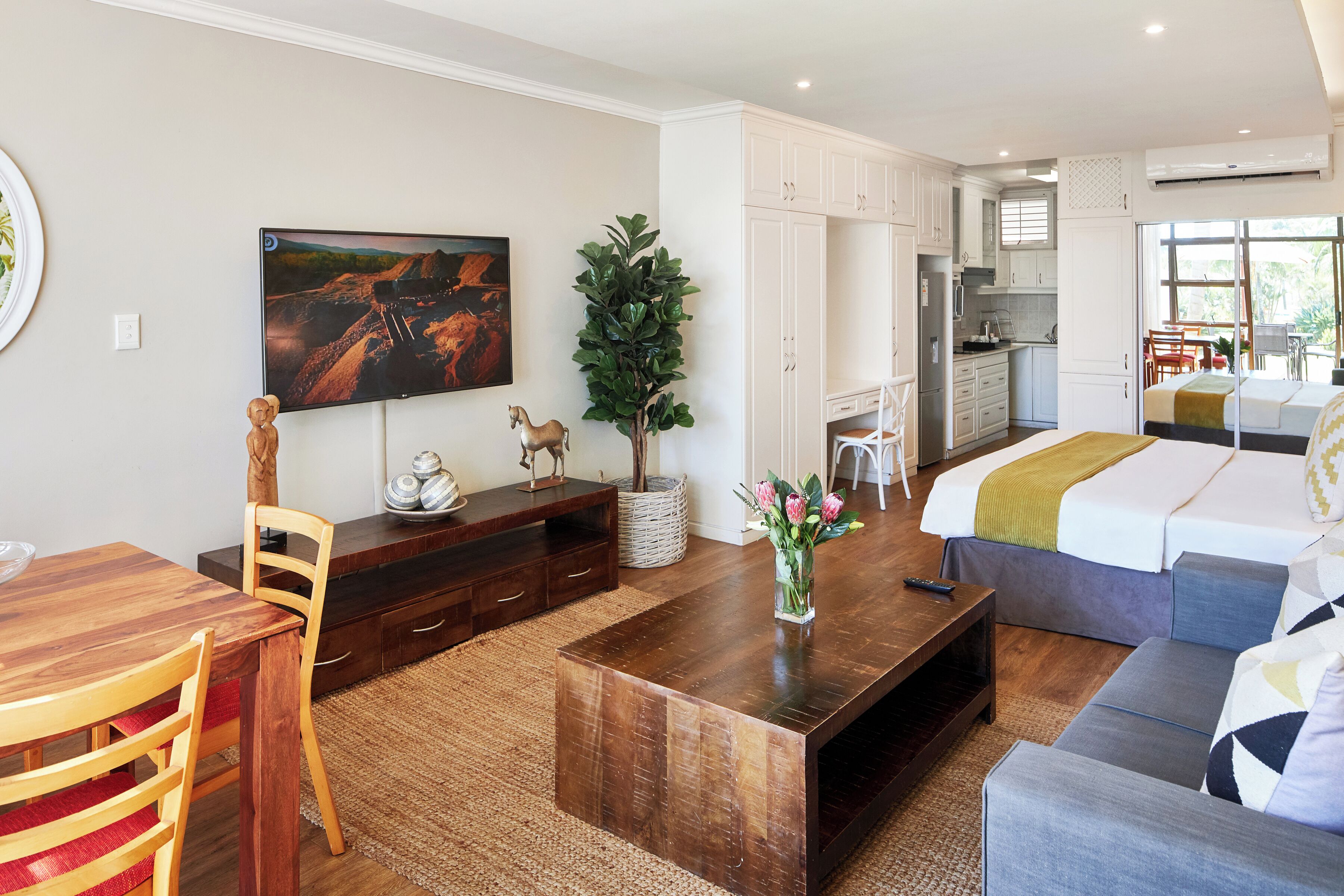 Een moderne, goed verlichte kamer met een houten eettafel, een bank, een bed met een groene deken, een televisie, en een zichtbare keuken door een open deur.