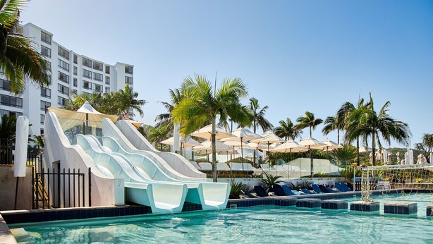 A swimming pool with a water slide, lounge chairs, palm trees, and a building in the background. -  First Group Breakers Resort (Umhlanga)