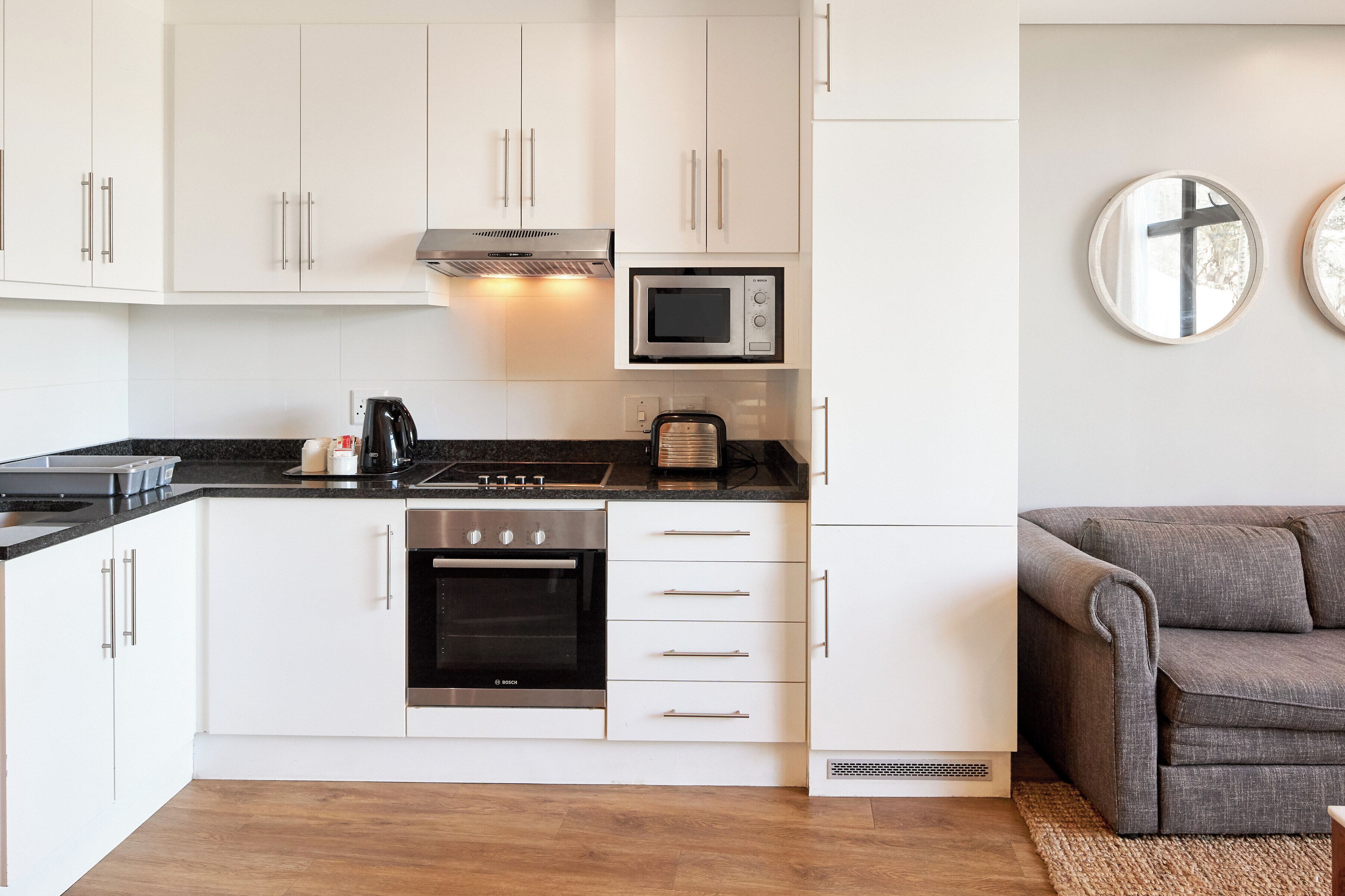 A modern kitchen with white cabinets, a built-in oven, a microwave, and a coffee maker. There is a gray sofa in the adjacent living area.