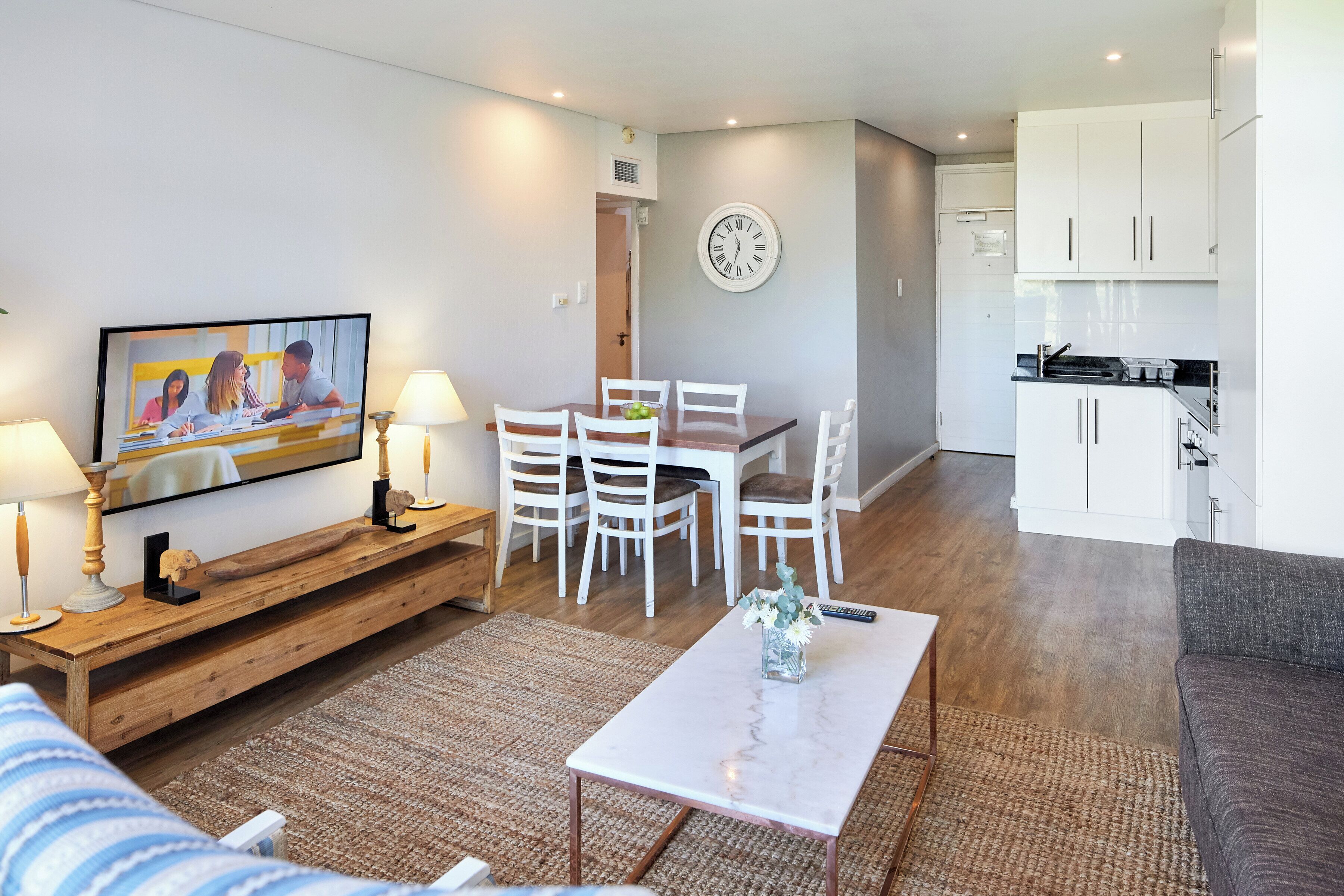 A modern living room with a TV, dining table, and chairs, connected to a kitchen area with white cabinets and a clock on the wall.