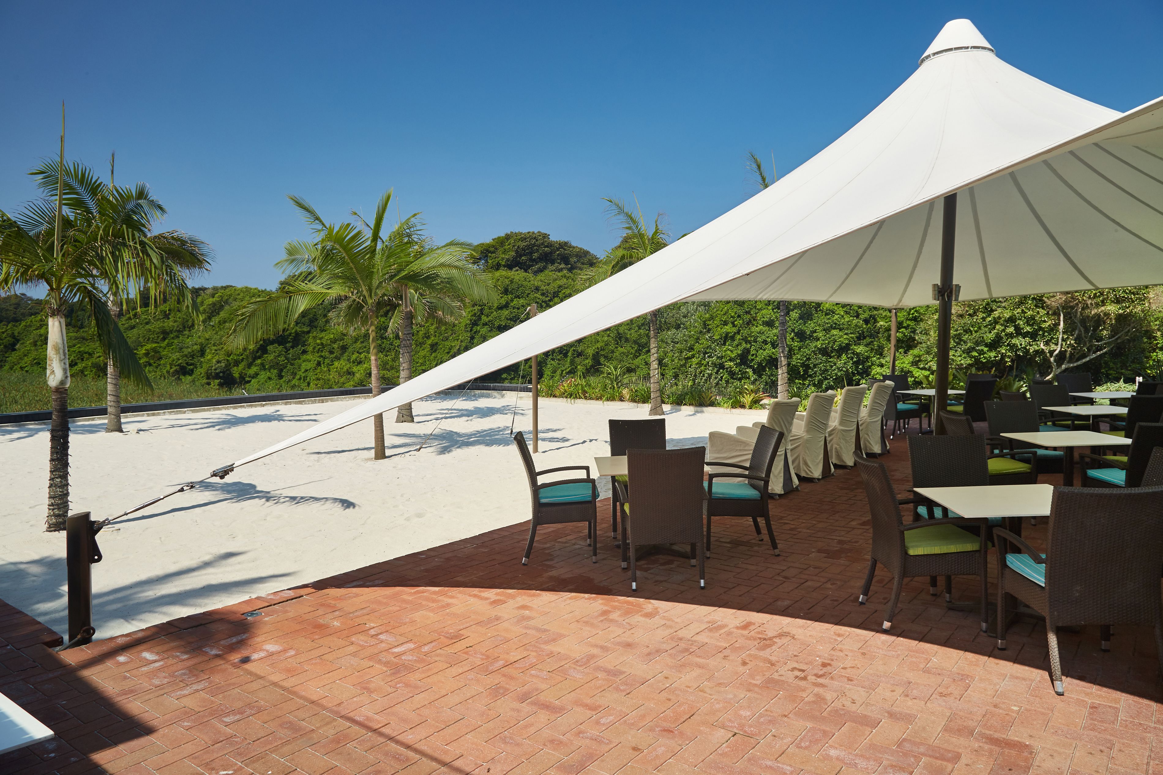 Un área de comedor al aire libre con mesas y sillas, un toldo blanco y vistas a la playa y palmeras.