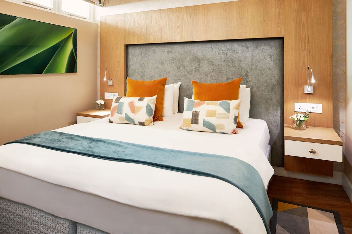 A modern hotel room with a large bed, wooden headboard, and a nightstand with a lamp.