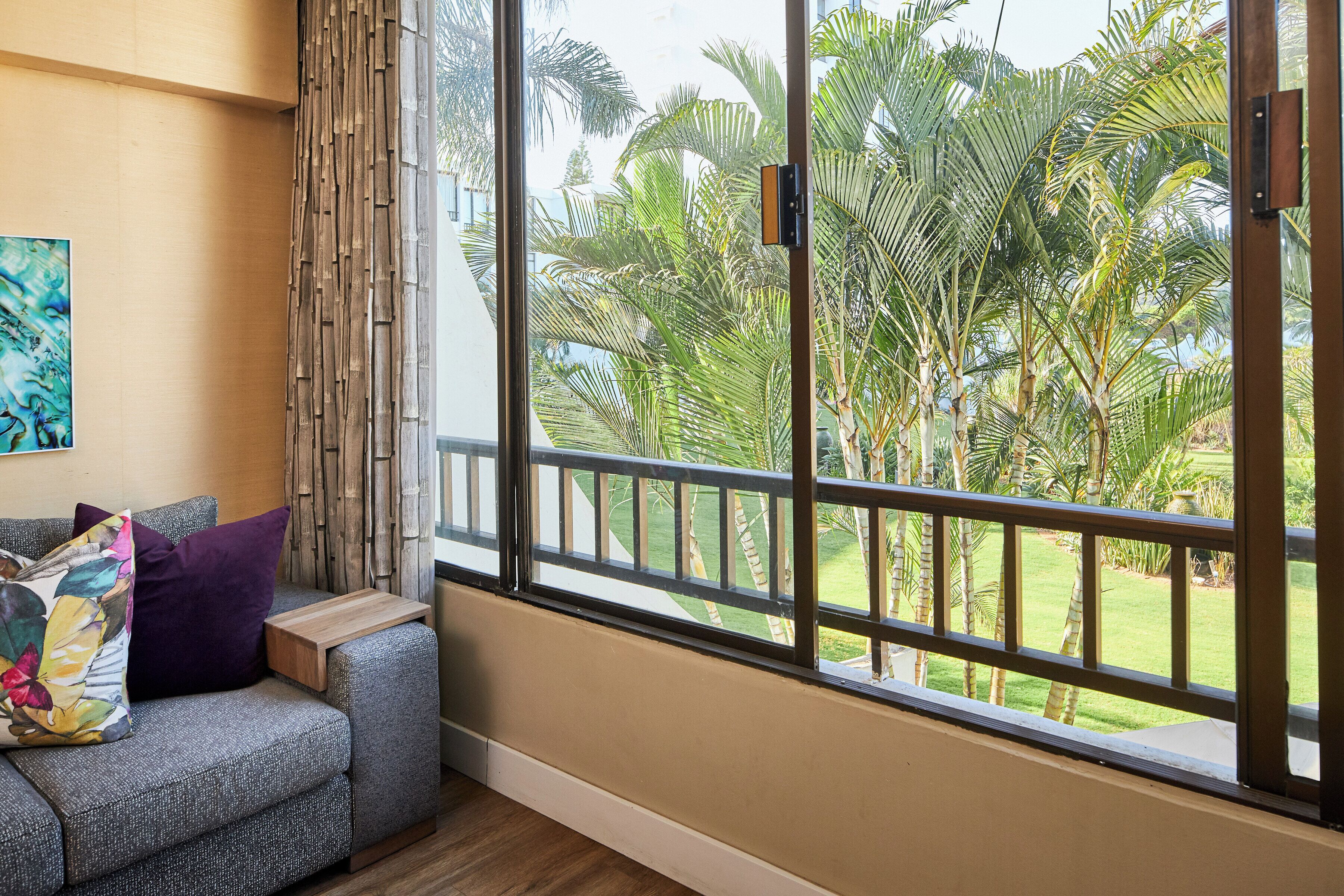 Una habitación con un ventanal grande, un sofá gris con cojines y vistas a palmeras.