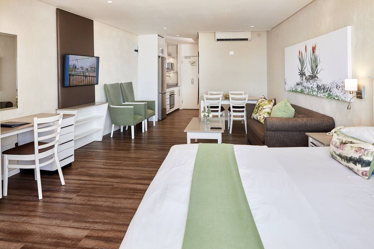 A hotel room with a bed, a desk, a chair, a sofa, a dining table, and a kitchen area.