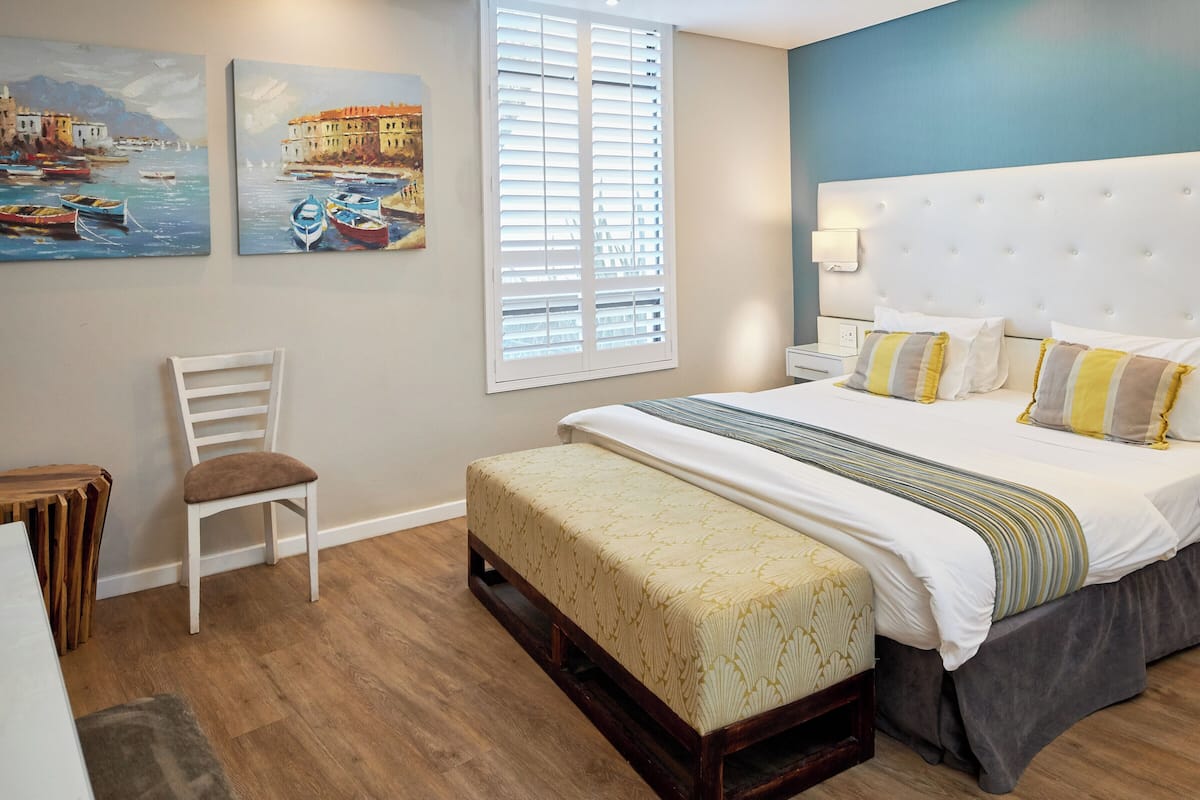A bedroom with a bed, a chair, a bench, a window with blinds, and two paintings on the wall.