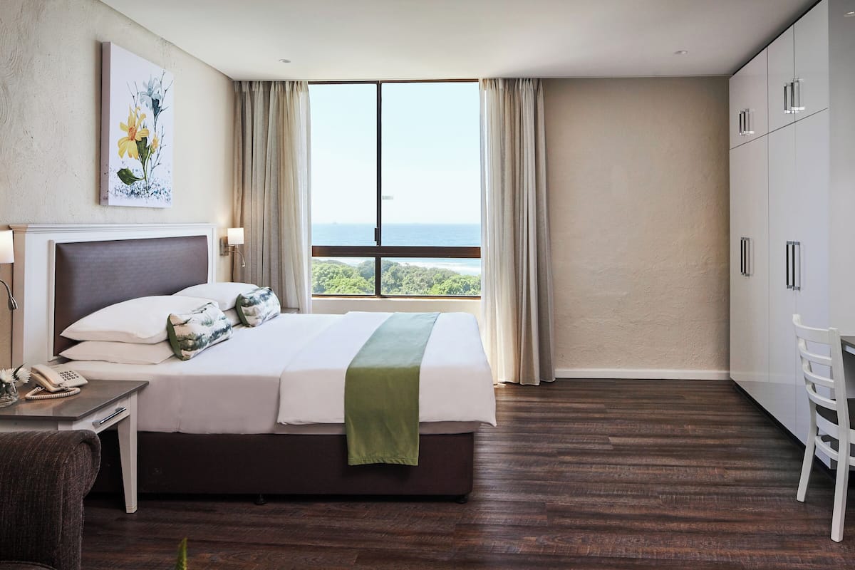 A hotel room with a large bed, a desk with a phone, a chair, and a view of the ocean through a large window.