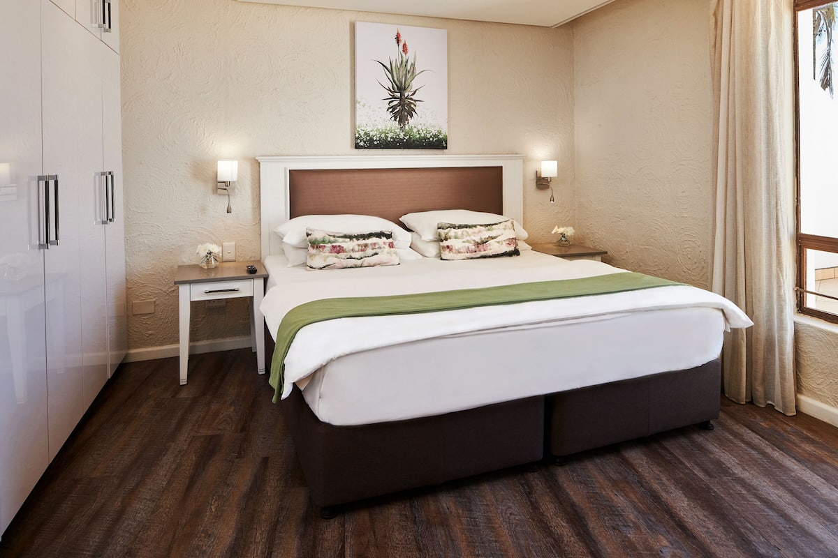 A hotel room with a large bed, bedside tables, a nightstand, a wall-mounted lamp, and a window with curtains.