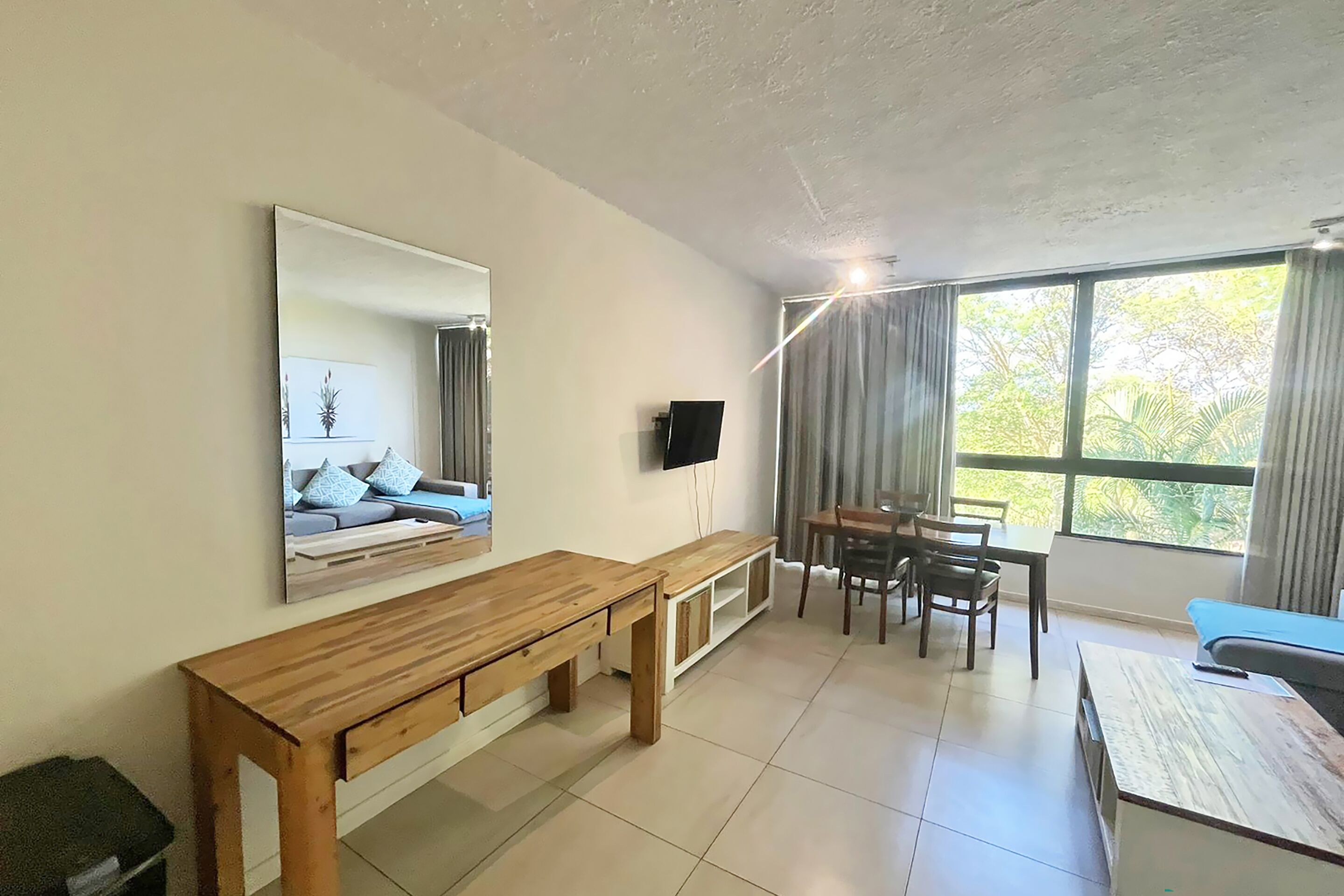 Een moderne woonkamer met een houten bankje, een eettafel met stoelen, een aan de muur bevestigde televisie en grote ramen met gordijnen.