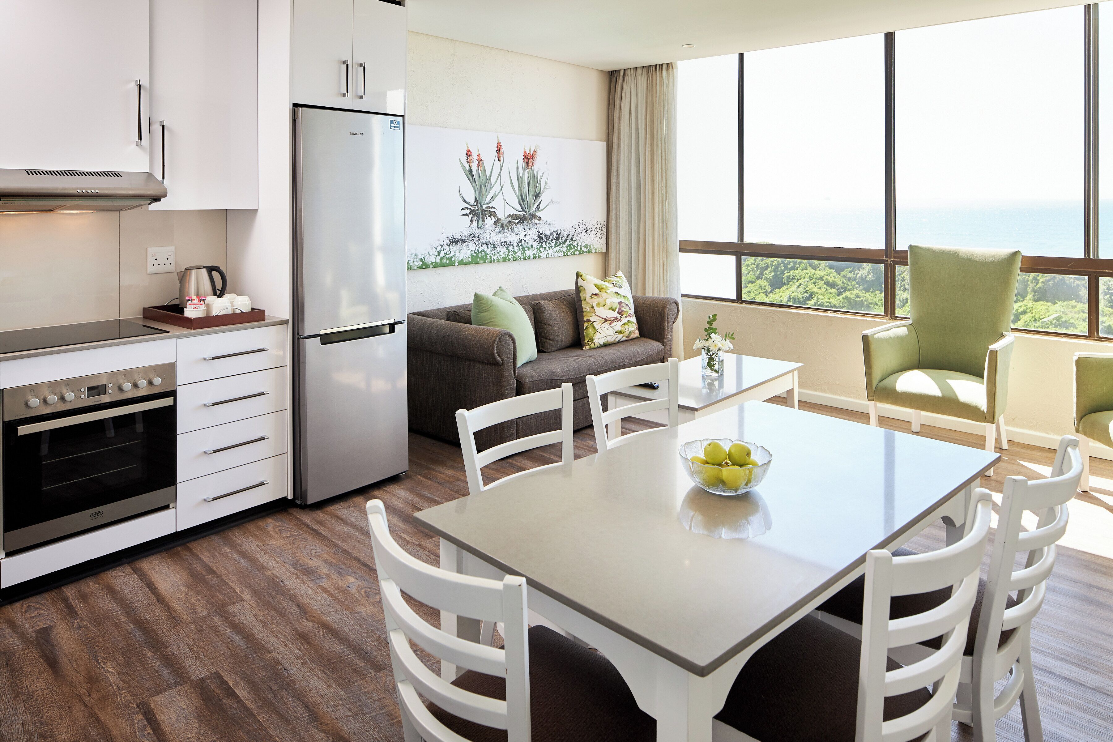 A modern kitchen with white cabinetry, a stainless steel refrigerator, and a built-in oven. There is a dining area with a table and chairs, and a large window offering a view of the ocean.