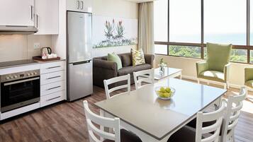 A modern kitchen with white cabinetry, a stainless steel refrigerator, and a built-in oven. There is a dining area with a table and chairs, and a large window offering a view of the ocean.