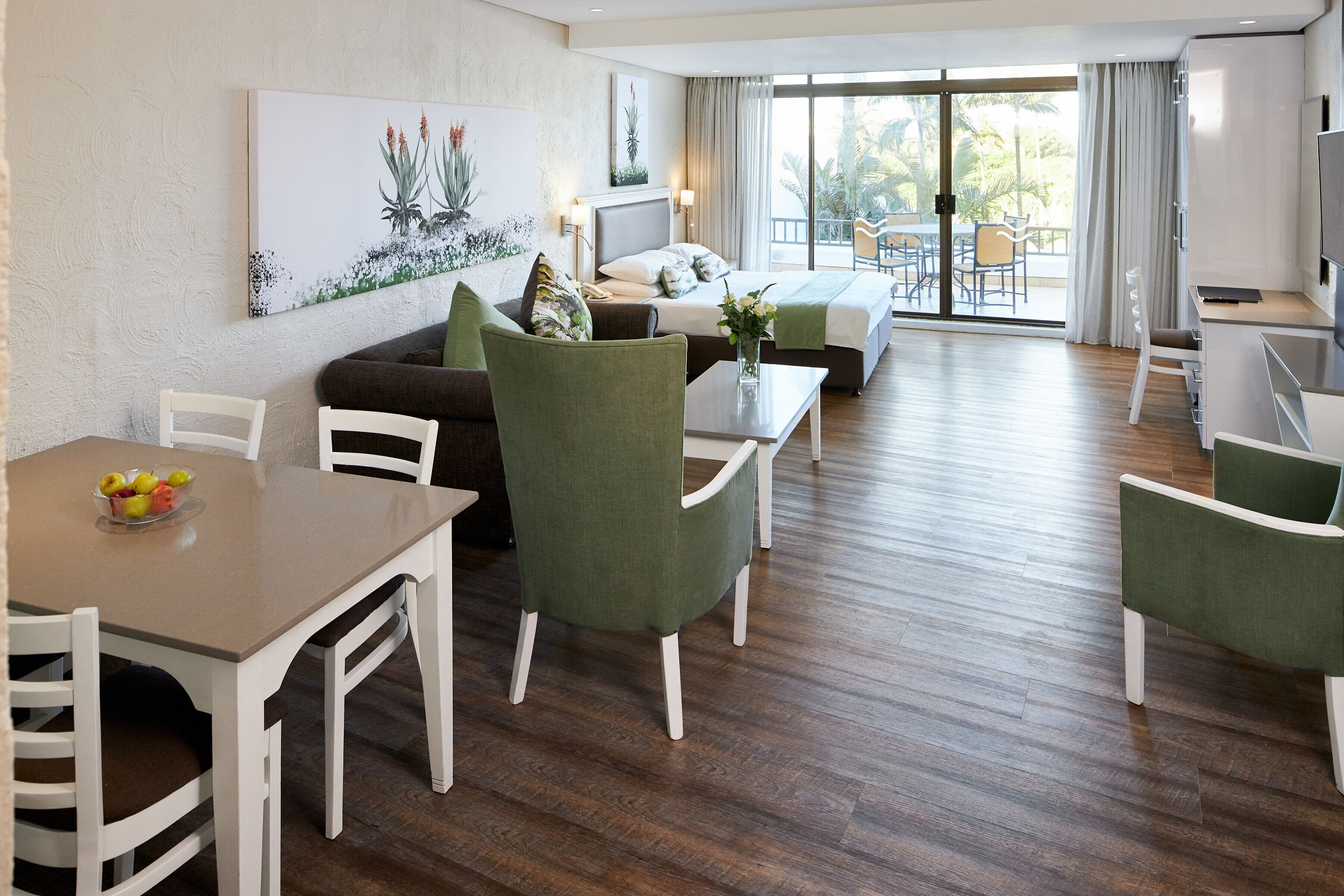 A hotel room with a bed, a dining area with a table and chairs, a balcony with a view of palm trees, and a TV on a stand.