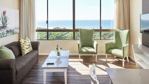 A modern living room with a large window offering a view of the ocean and greenery. There is a sofa, two armchairs, and a coffee table with a remote control on it. -  First Group Breakers Resort (Umhlanga)