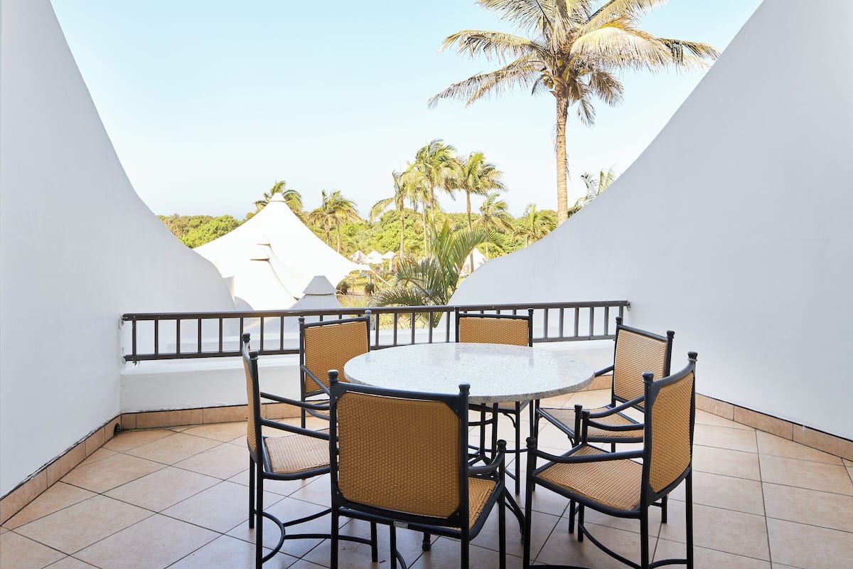 A balcony with a round table and wicker chairs, overlooking a beach with palm trees and a white building.