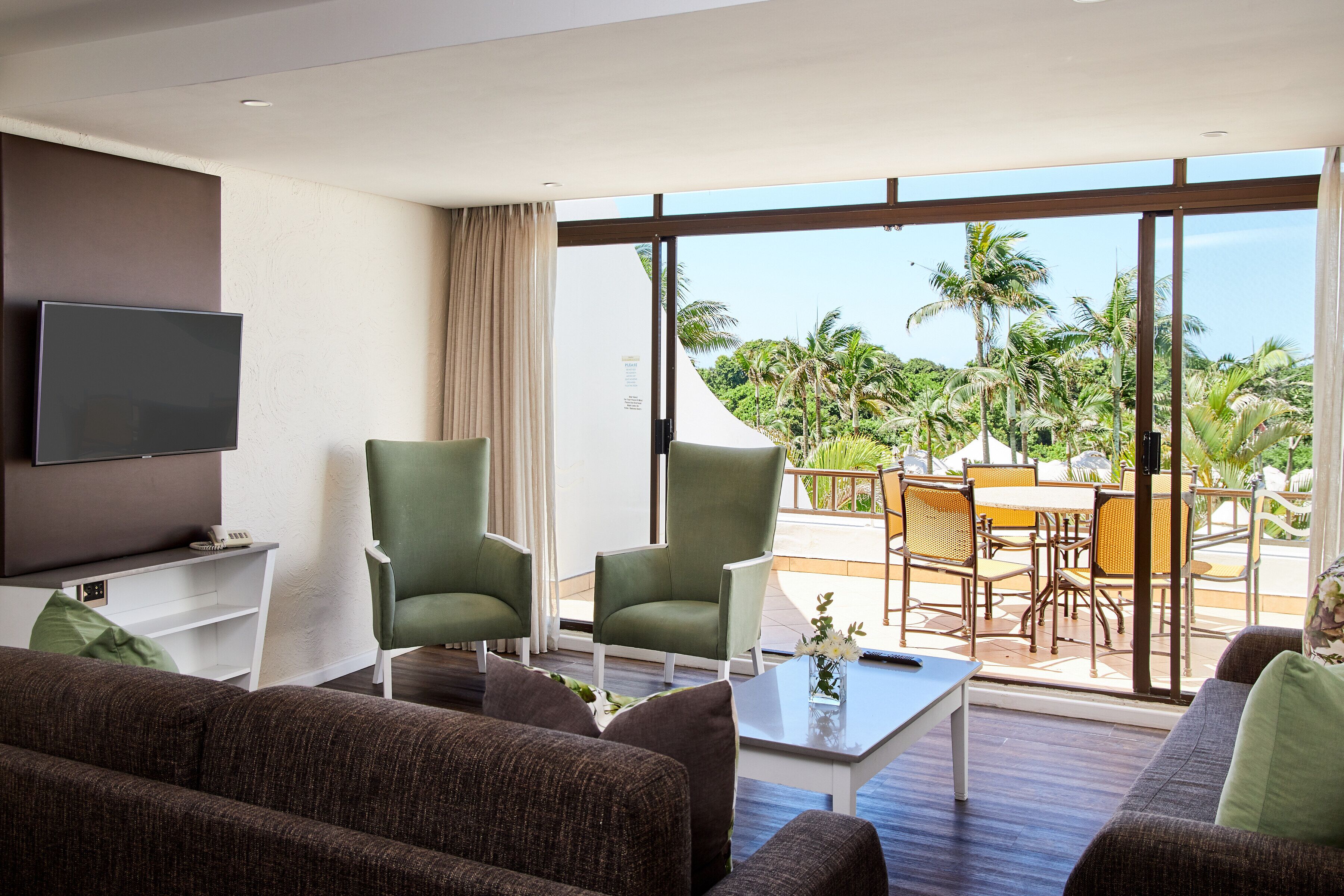 Una sala de estar moderna con una televisión de pantalla plana, sillones verdes, un sofá y una mesa de centro. Se ve un balcón con muebles de exterior y palmeras a través de ventanas amplias.