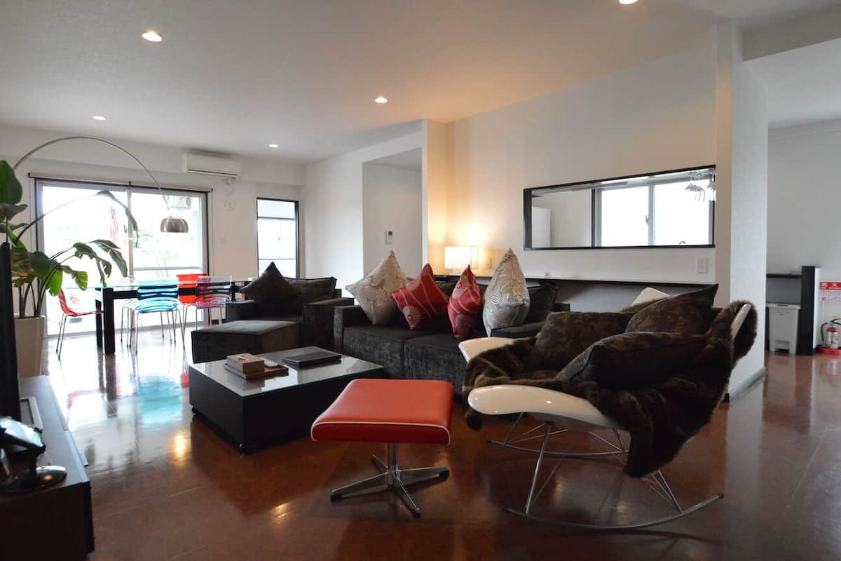 Una sala de estar moderna con sofá, reposapiés y sillones, un comedor con sillas y una zona de cocina.