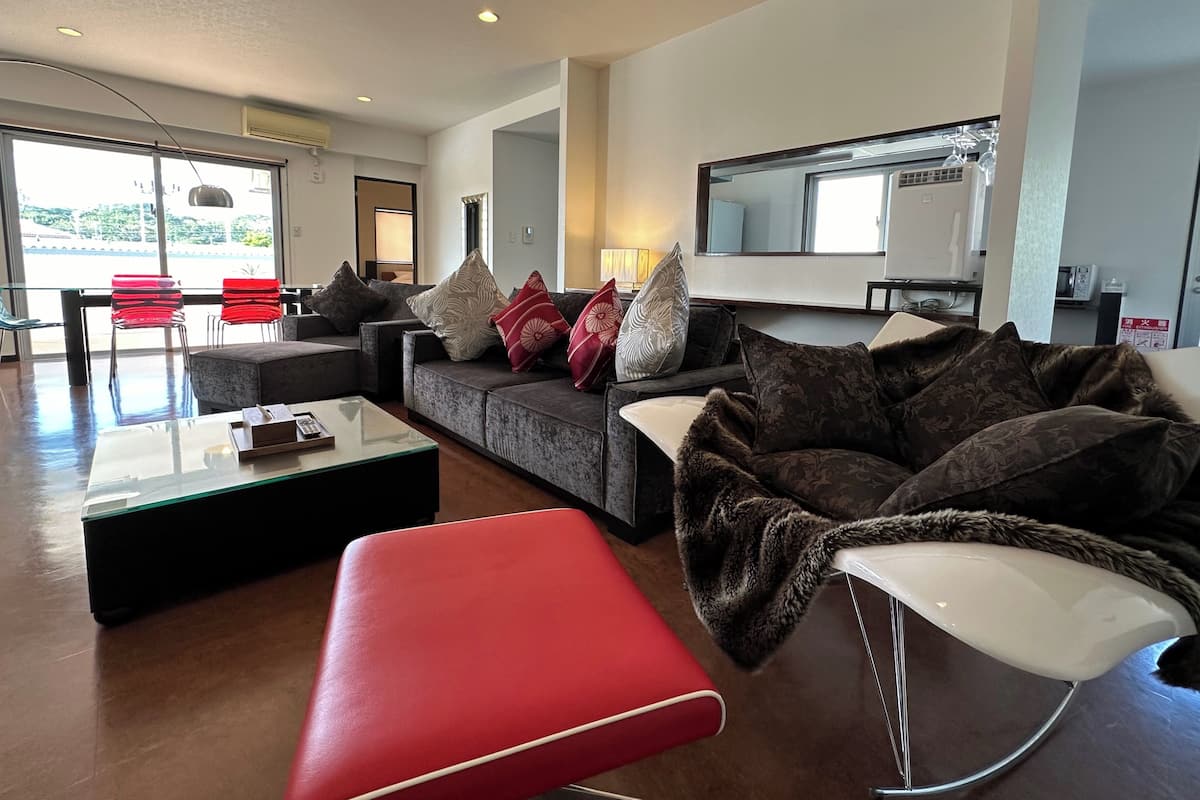 Amplia sala de estar con un sofá grande, un reposapiés rojo y una mesa de centro de vidrio. Hay un televisor montado en la pared y una ventana que ofrece vistas al exterior.