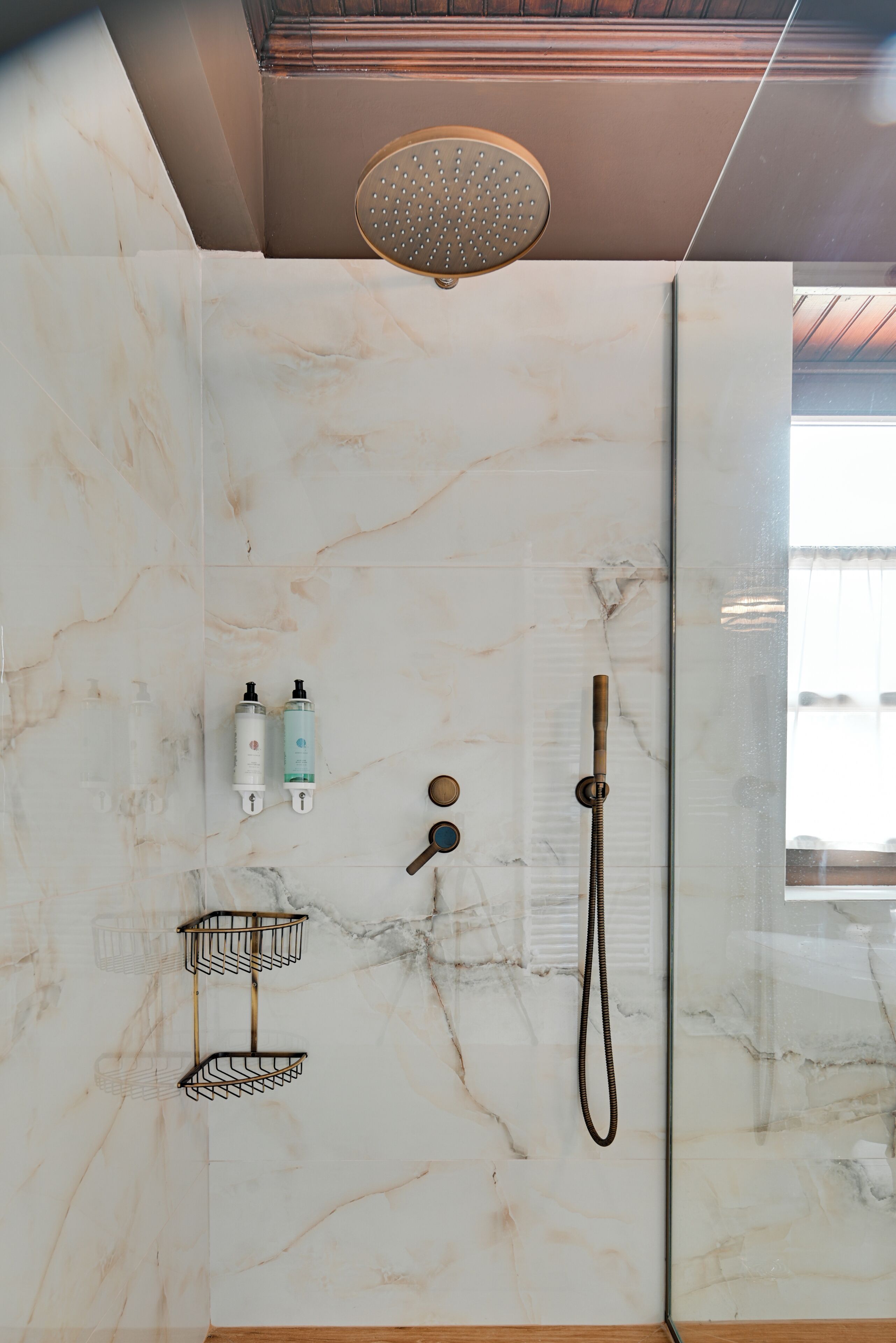 A bathroom with a shower, marble walls, a handheld showerhead, a handheld shower attachment, and two bottles of shampoo.