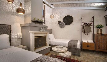 Habitación de hotel compacta con cama, mesita de noche y una pequeña mesa redonda. Cuenta con una chimenea, un estante en la pared con adornos y un perchero.