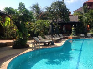 A swimming pool with lounge chairs and umbrellas, surrounded by lush greenery and a building with a tiled roof.
