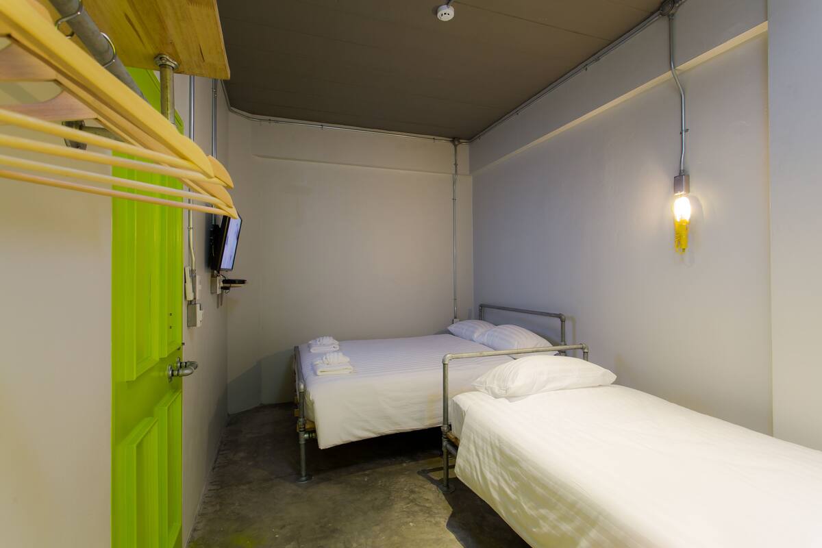 A small, single-room accommodation with two beds, a green door, and a wall-mounted light fixture.