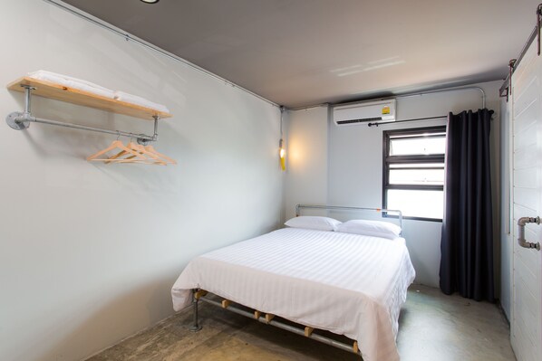 A small, single-bed room with a metal bed frame, a white bedspread, a wooden shelf with hangers, a wall-mounted light, and an air conditioning unit.