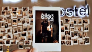 Una mano tiene una foto Polaroid con la scritta 'bloo hostel' di fronte a un muro ricoperto da numerose piccole fotografie e la parola 'bloo' scritta in grandi caratteri.