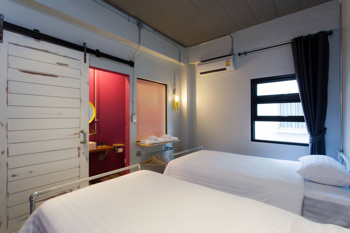 A hotel room with two beds, a red door, a window with curtains, and an air conditioning unit.