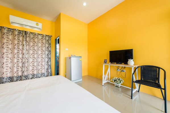 Une chambre d’hôtel avec un lit, un climatiseur blanc, un réfrigérateur, un téléviseur posé sur un meuble blanc et une chaise noire.