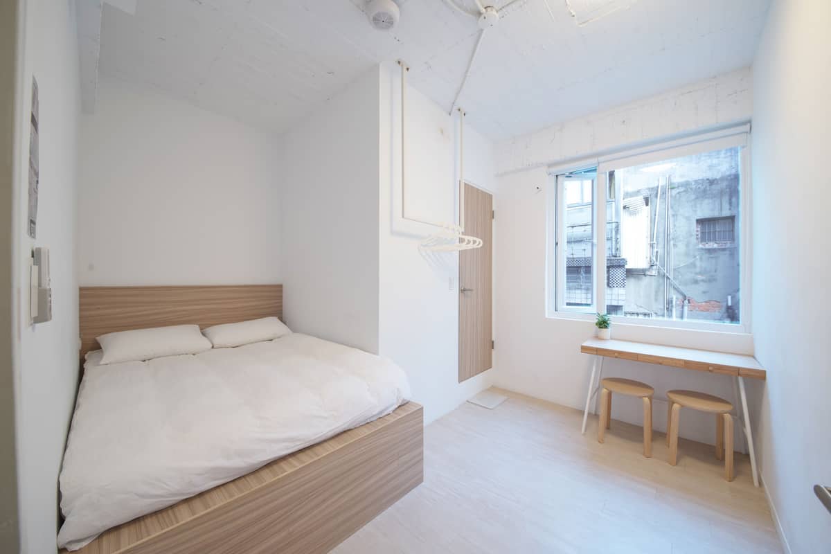Une chambre moderne avec un lit en bois, un bureau et des tabourets, une fenêtre donnant sur des immeubles et un luminaire suspendu.
