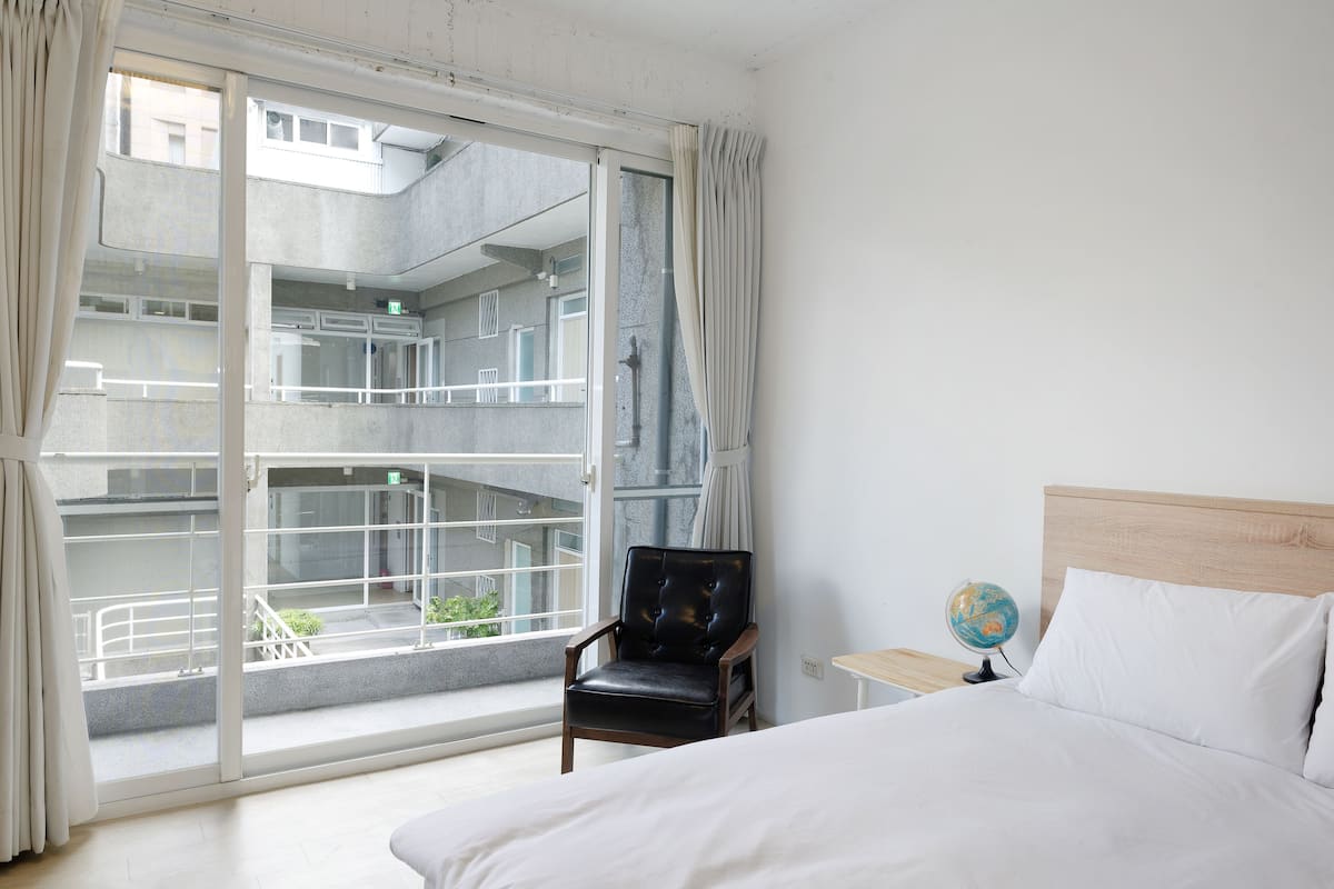 Une chambre d’hôtel avec une grande fenêtre, un lit, une chaise et une table de chevet sur laquelle se trouve un globe terrestre.
