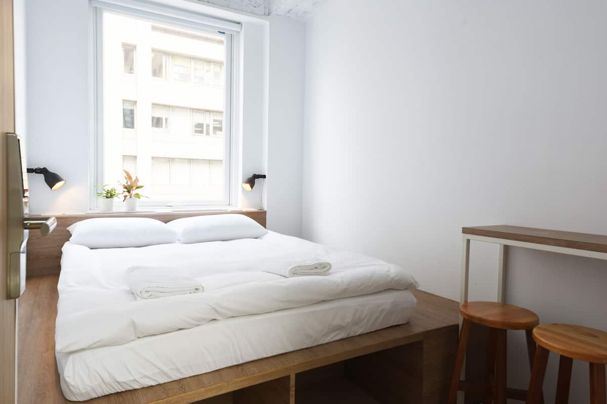 Un lit bien fait, avec du linge de lit blanc, un banc en bois au pied du lit et deux lampes fixées au mur.