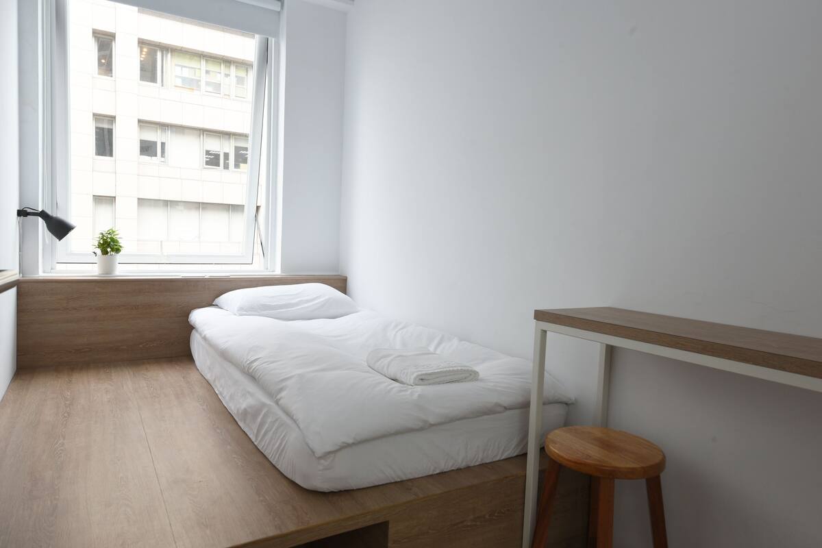 Une chambre à coucher minimaliste comprenant un lit, un petit tabouret, une table de chevet, une plante en pot et une fenêtre donnant sur un immeuble.
