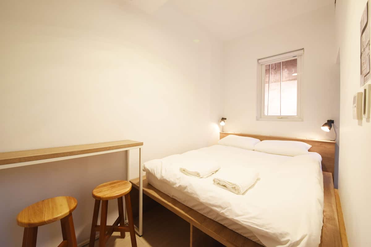 Une chambre avec un lit simple, deux tabourets en bois, une fenêtre avec des rideaux et des lampes fixées au mur.