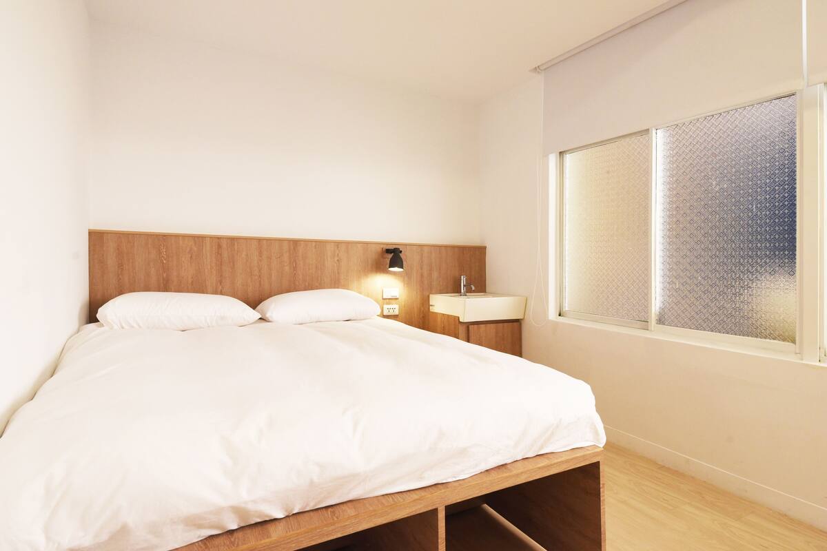 Une chambre à coucher avec une tête de lit en bois, une table de chevet avec une lampe, une fenêtre à vitres dépolies et un couvre-lit blanc.