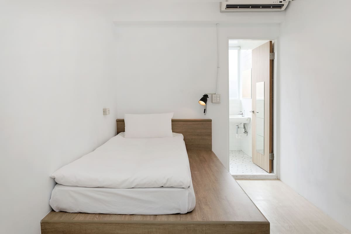 Une chambre à coucher au style minimaliste, avec un lit surélevé en bois, du linge de lit blanc, une lampe de chevet et une vue sur une salle de bain dotée d’une douche en verre.