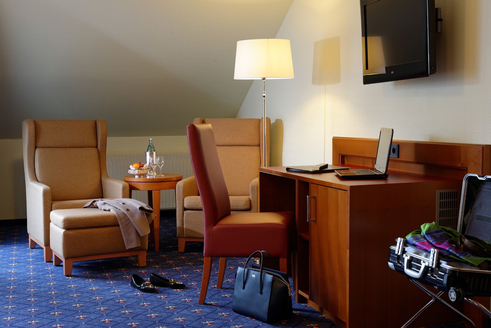 Ein Hotelzimmer mit zwei Sesseln, einem Schreibtisch mit Laptop, einer Lampe, einem Fernseher und einem Koffer.