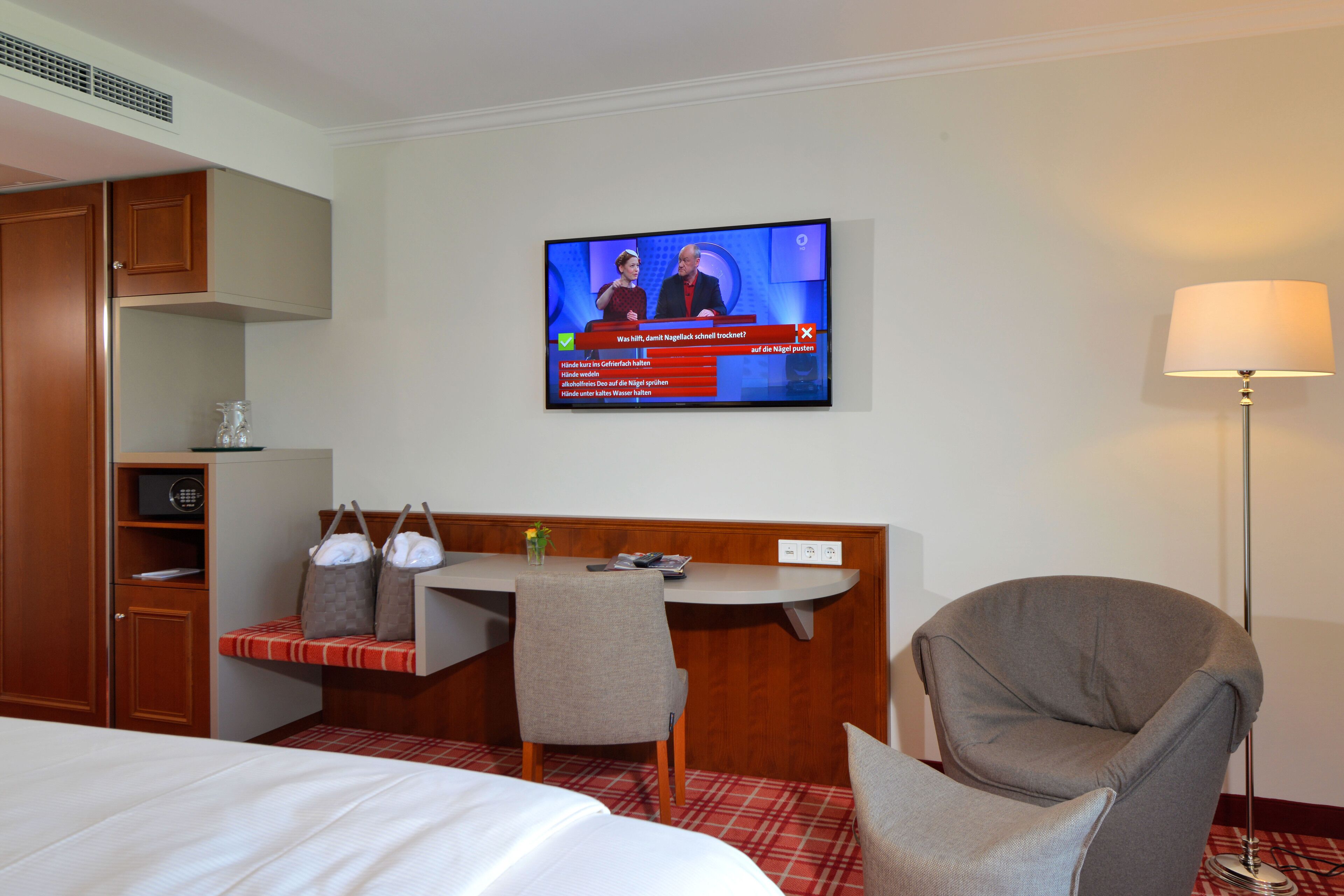 Ein Hotelzimmer mit Bett, Schreibtisch mit Stuhl, Fernseher der eine Nachrichtensendung zeigt und einer Lampe.