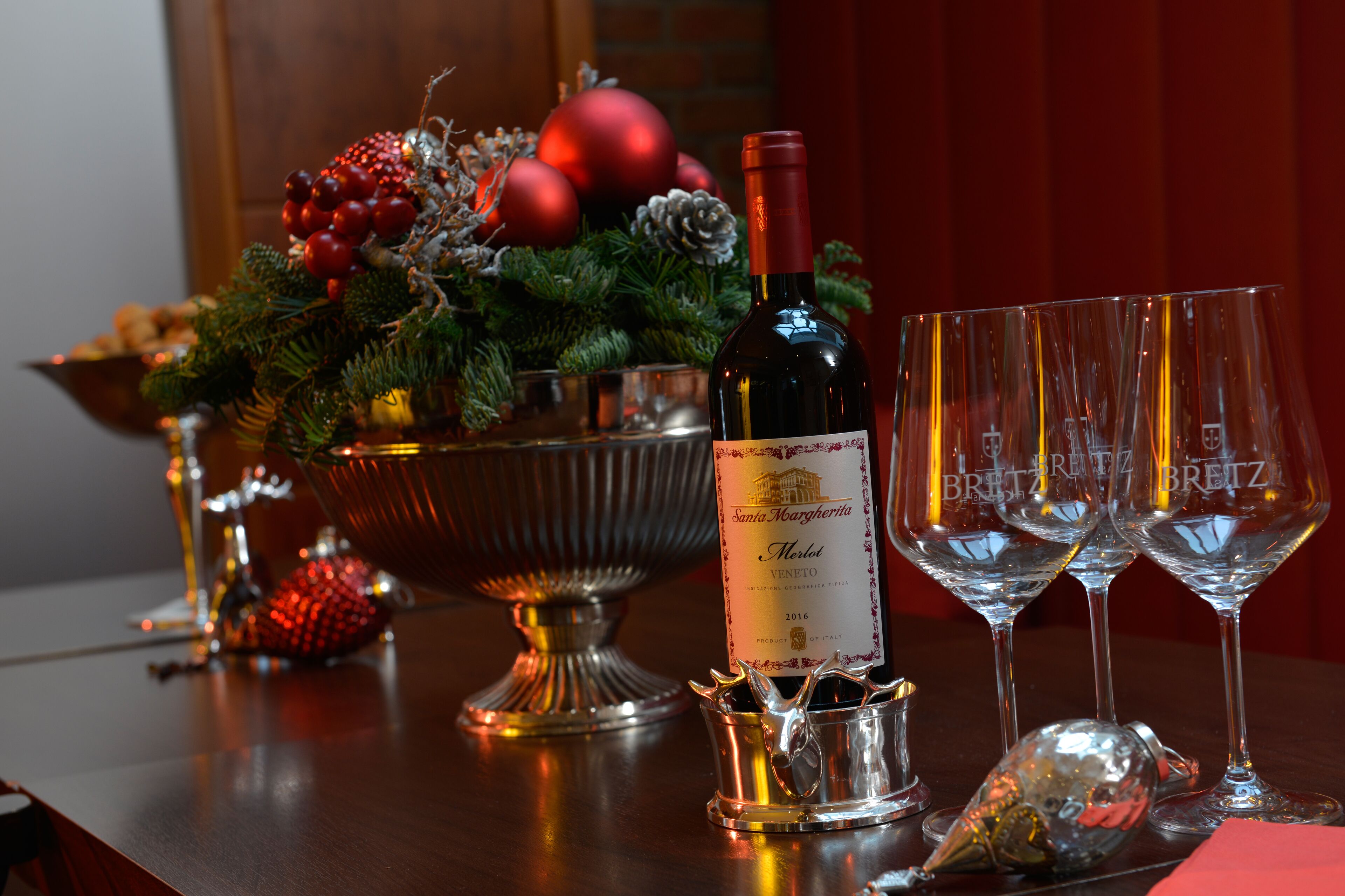 A table with a bottle of wine, two glasses, a silver bowl with Christmas decorations, and a small metal pitcher.