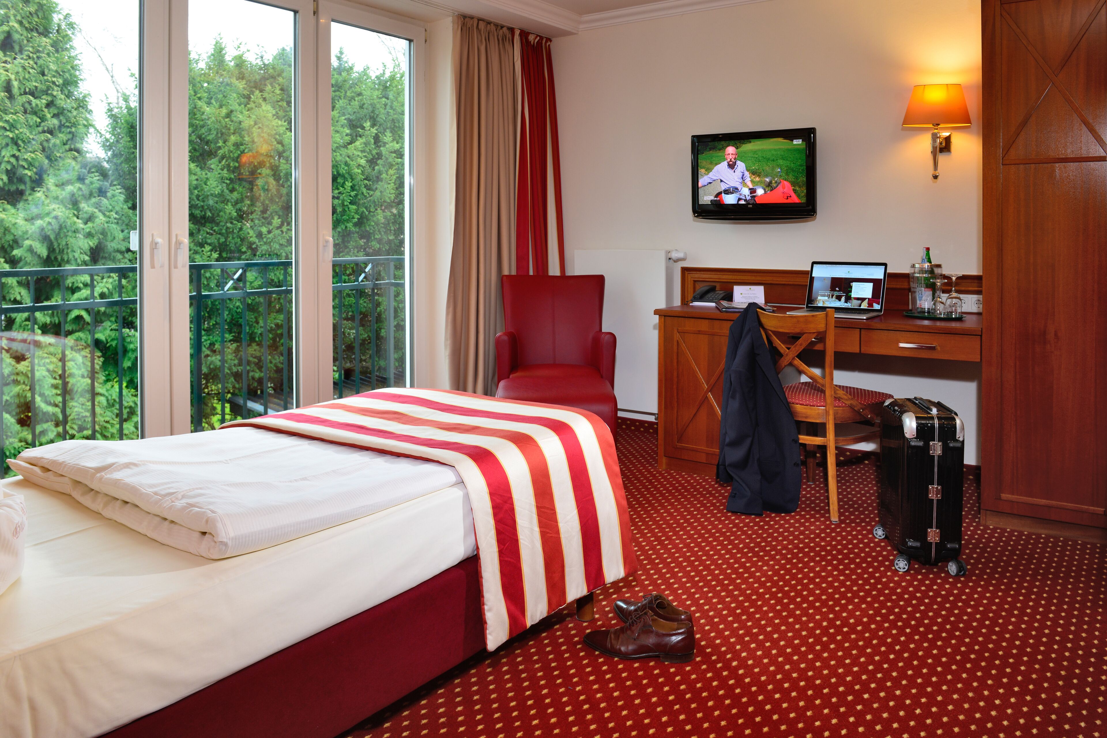 A hotel room with a bed, a desk with a laptop, a chair, a suitcase, and a TV on the wall.
