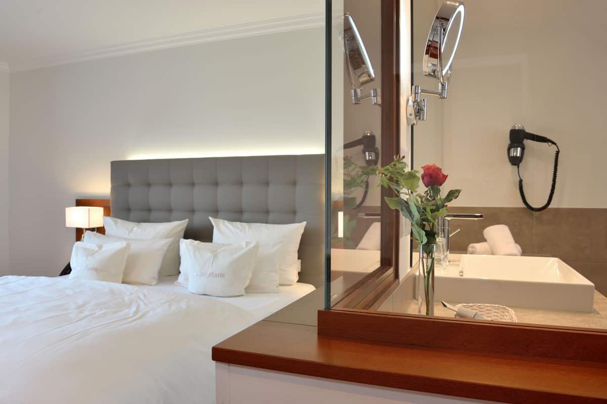 Quarto de hotel com uma cama grande, abajur, espelho refletindo um banheiro com pia e porta-toalhas.