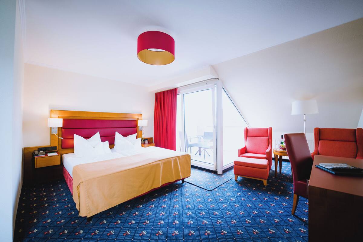 Quarto de hotel com cama, escrivaninha, cadeira e uma janela grande.